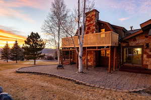 Back of house at dusk with stone siding, a gambrel roof, a patio, a wooden deck, and a yard