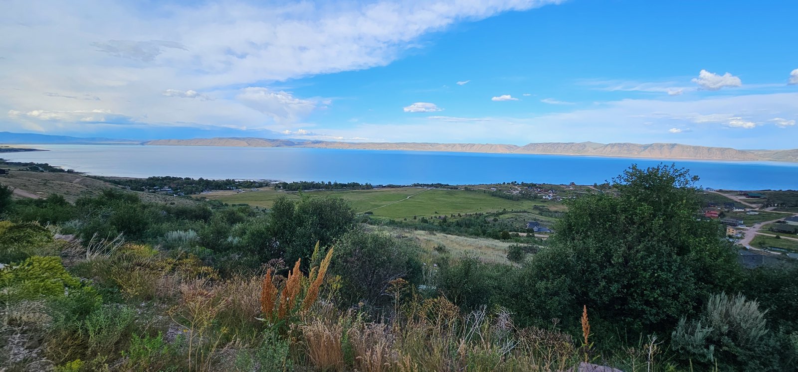 Expansive water body with distant mountain ranges under a blue sky, featuring a varied landscape of green fields and natural foliage
