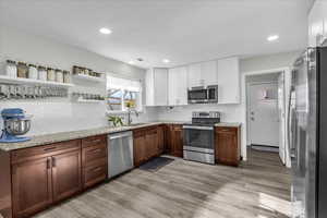 Two tone kitchen featuring open shelves, stainless steel appliances, two tone cabinets, light stone countertops, and decorative backsplash