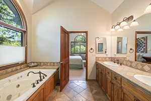 Ensuite bathroom featuring vaulted ceiling, double vanity, backsplash, stone tile flooring, and a tub with jets