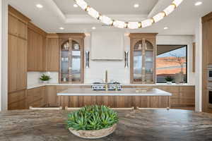 Kitchen with glass fronted cabinets, an island with sink, ventilation hood, light stone counters, and recessed lighting