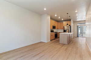 Kitchen featuring light wood finish cabinetry, stainless steel appliances, decorative light fixtures, an island with sink, and light wood-style floors