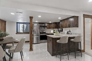 Kitchen with dark wood finish cabinetry, a peninsula, stainless steel appliances, a breakfast bar area, and light stone counters
