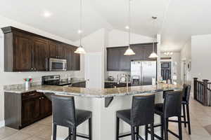 Kitchen with light tile patterned floors, stainless steel appliances, light stone counters, dark wood finish cabinetry, and lofted ceiling