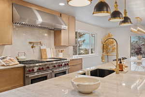 Kitchen with hood vent, light stone counters, double oven Wolf range, hanging light fixtures, and decorative backsplash