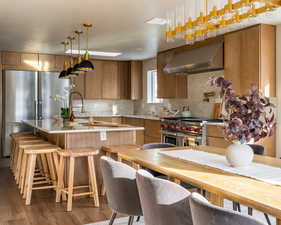 Kitchen featuring range hood, stainless steel appliances, decorative backsplash, dark wood-style flooring, and pendant lighting