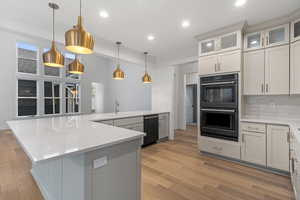 Kitchen with pendant lighting, black appliances, light wood-style floors, light stone counters, and glass fronted cabinets