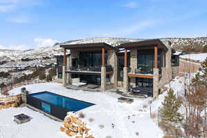 Snow covered house with stone siding, a patio, an outdoor pool, a mountain view, and a balcony