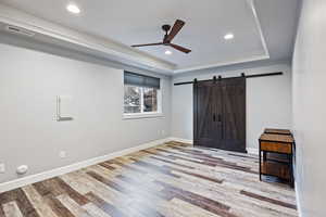 Unfurnished bedroom featuring a barn door, ceiling fan, light wood finished floors, recessed lighting, and a tray ceiling