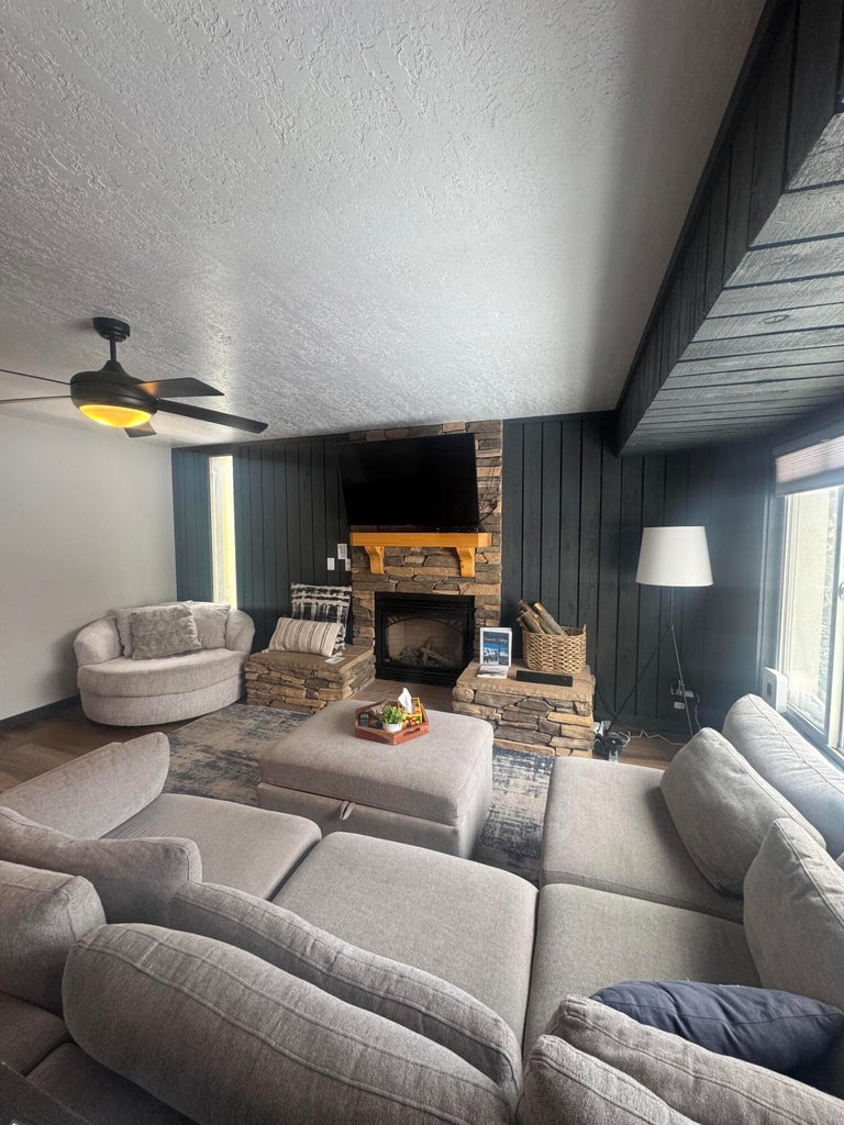 Living room featuring a stone fireplace, wood walls, a textured ceiling, ceiling fan, and wood finished floors