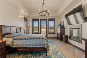 Bedroom with a raised ceiling, light stone finish floors, a glass covered fireplace, suspended lighting, and access to outside