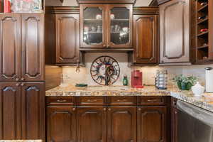 Kitchen featuring stainless steel dishwasher, glass insert cabinets, dark wood finish cabinets, and light stone counters