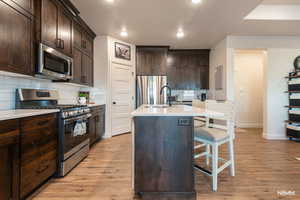 Kitchen featuring dark wood finish cabinetry, stainless steel appliances, tasteful backsplash, light wood-style flooring, and a center island with sink