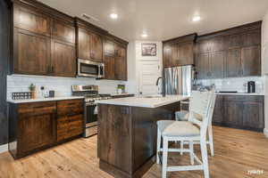 Kitchen with dark wood finish cabinets, stainless steel appliances, tasteful backsplash, a breakfast bar, and a center island with sink