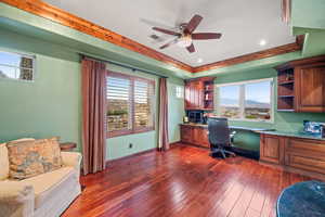 Home office with built in desk, a raised ceiling, healthy amount of natural light, a ceiling fan, and dark wood-type flooring