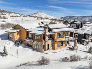 Snow covered house with stone siding, a balcony, a mountain view, and a chimney