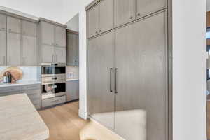 Kitchen with gray cabinets, light wood-type flooring, backsplash, double oven, and light stone countertops