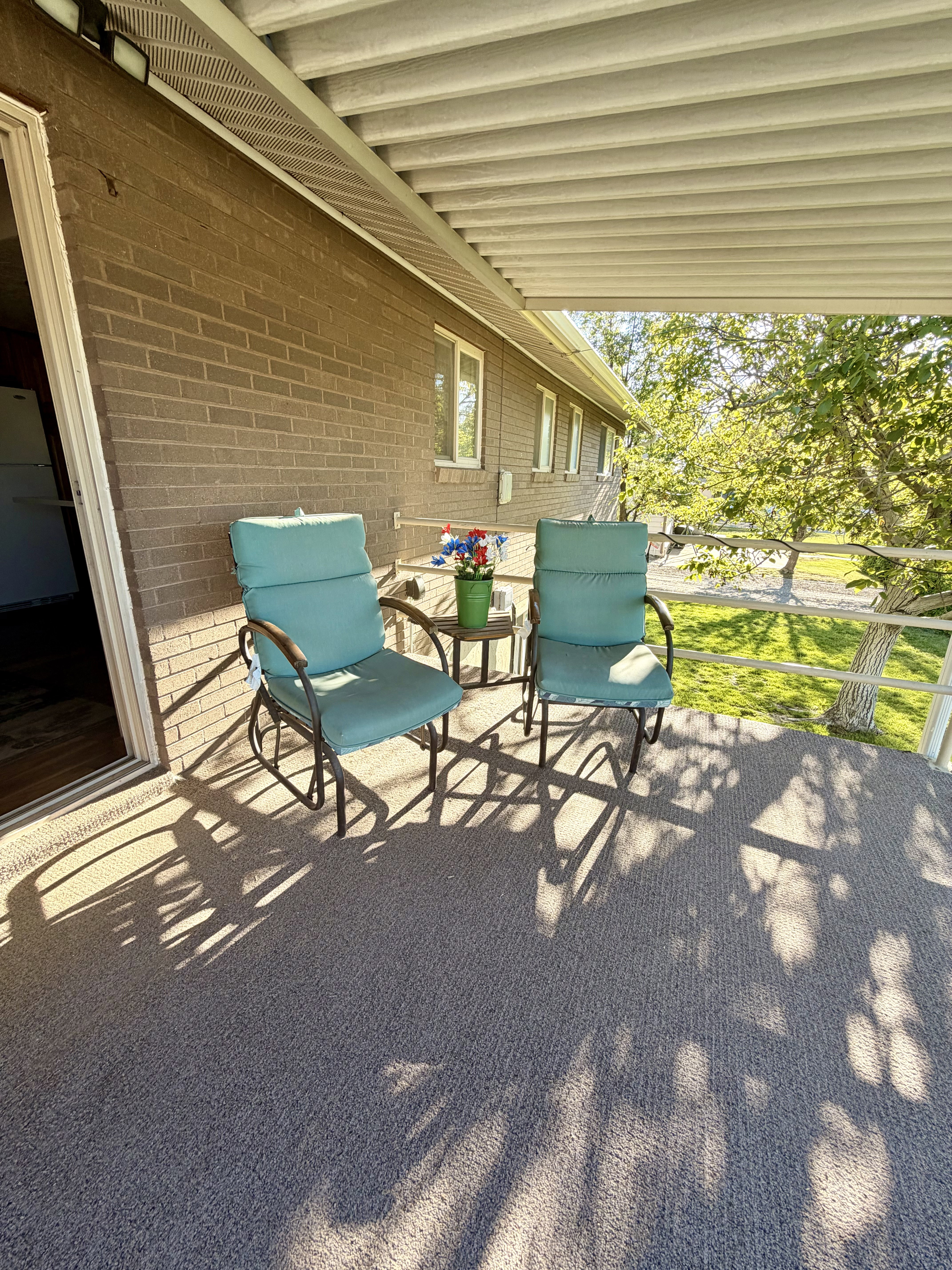 Covered outdoor patio featuring a brick exterior, textured outdoor carpeting, white railing, and an overhead slatted roof