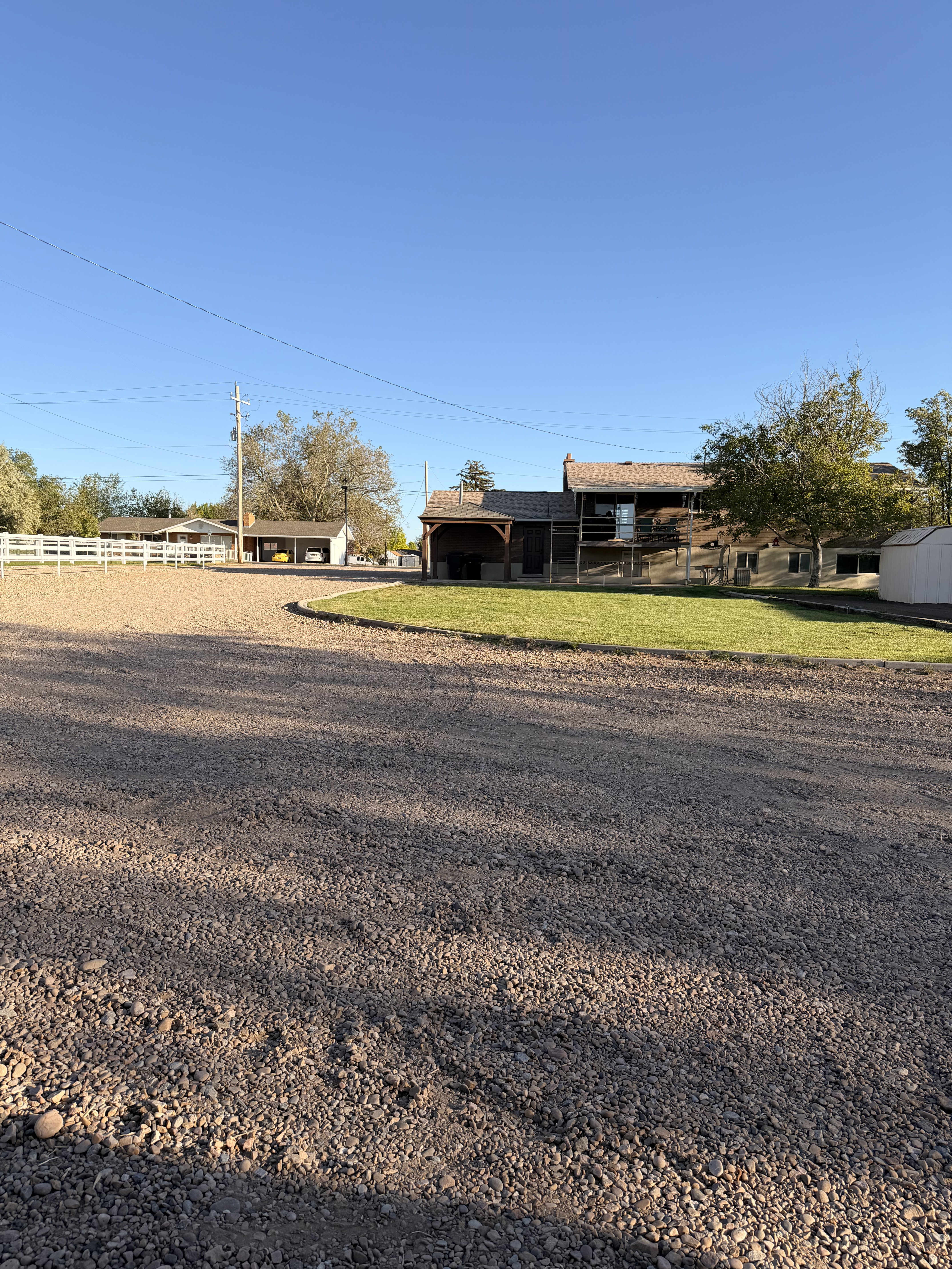 Expansive gravel driveway leading to a multi-structure property featuring a white picket fence, a dedicated grassy area, and mature trees
