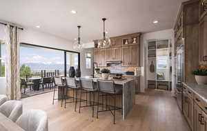 Kitchen featuring wood finish cabinetry, light wood-type flooring, a kitchen island with sink, decorative light fixtures, and light stone counters