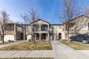 View of front of property featuring stone siding, a balcony, driveway, stucco siding, and a front yard