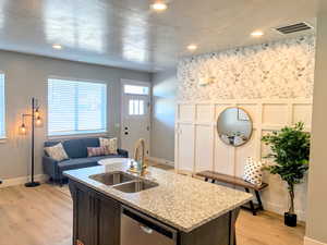 Kitchen with a decorative wall, stainless steel dishwasher, an island with sink, light wood finished floors, and light stone counters