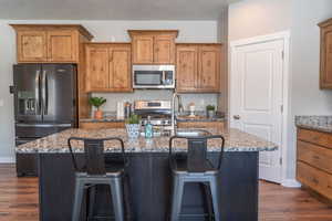 Kitchen with stainless steel appliances, light stone counters, an island with sink, wood finish cabinets, and a kitchen bar