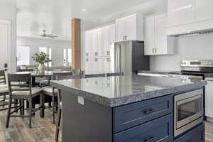 Kitchen with two tone cabinetry, stainless steel appliances, ceiling fan, light wood-style floors, and dark stone counters