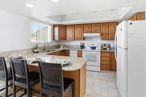 Kitchen featuring wood finish cabinetry, white appliances, light stone counters, a peninsula, and a raised ceiling