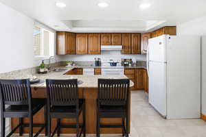 Kitchen featuring wood finish cabinets, white appliances, light stone countertops, a raised ceiling, and a peninsula
