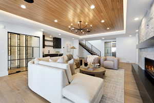 Living room with a wood tray ceiling, light wood-type flooring, a stone fireplace, bar with sink, and a chandelier