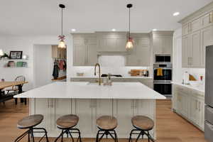 Kitchen featuring pendant lighting, stainless steel appliances, light wood-type flooring, light stone countertops, and an island with sink