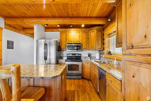Kitchen featuring stainless steel appliances, wood finish cabinetry, a kitchen bar, light stone counters, and a wood ceiling with exposed beams