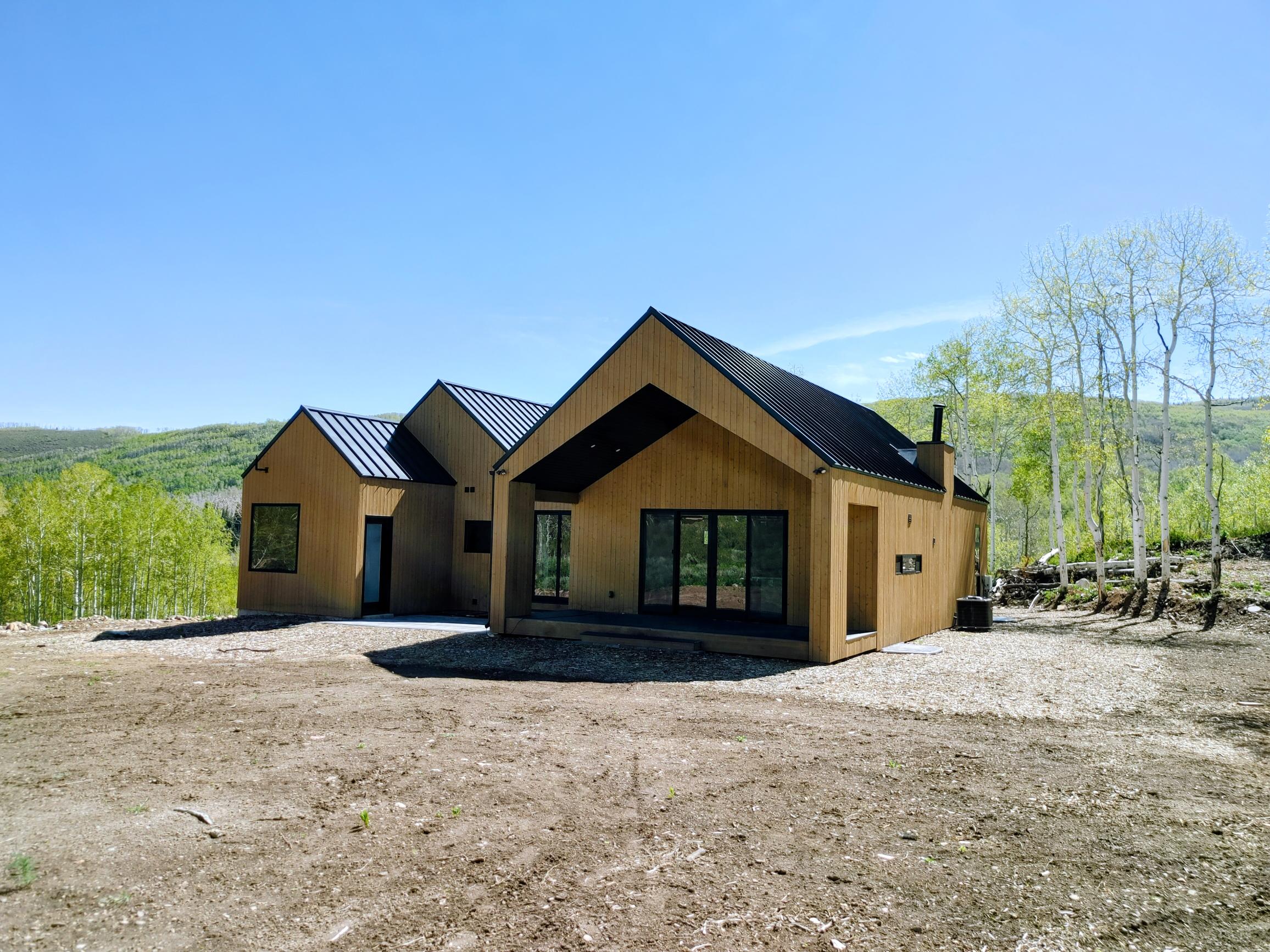 Contemporary home featuring a light wood-clad exterior, contrasting dark metal roofing, multiple large windows, and a black-framed multi-panel sliding glass door