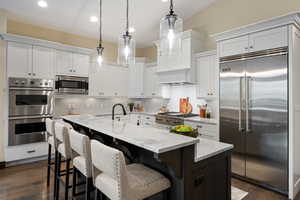 Kitchen featuring built in appliances, dual tone cabinetry, a center island with sink, pendant lighting, and light stone counters