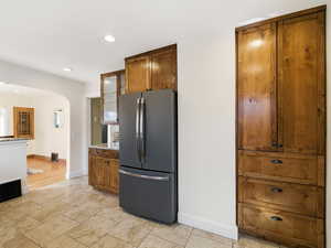 Kitchen featuring wood finish cabinets, glass insert cabinets, freestanding refrigerator, arched walkways, and light stone counters