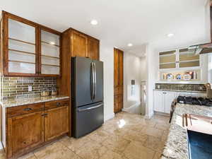 Kitchen with glass fronted cabinets, light stone countertops, backsplash, freestanding refrigerator, and range hood