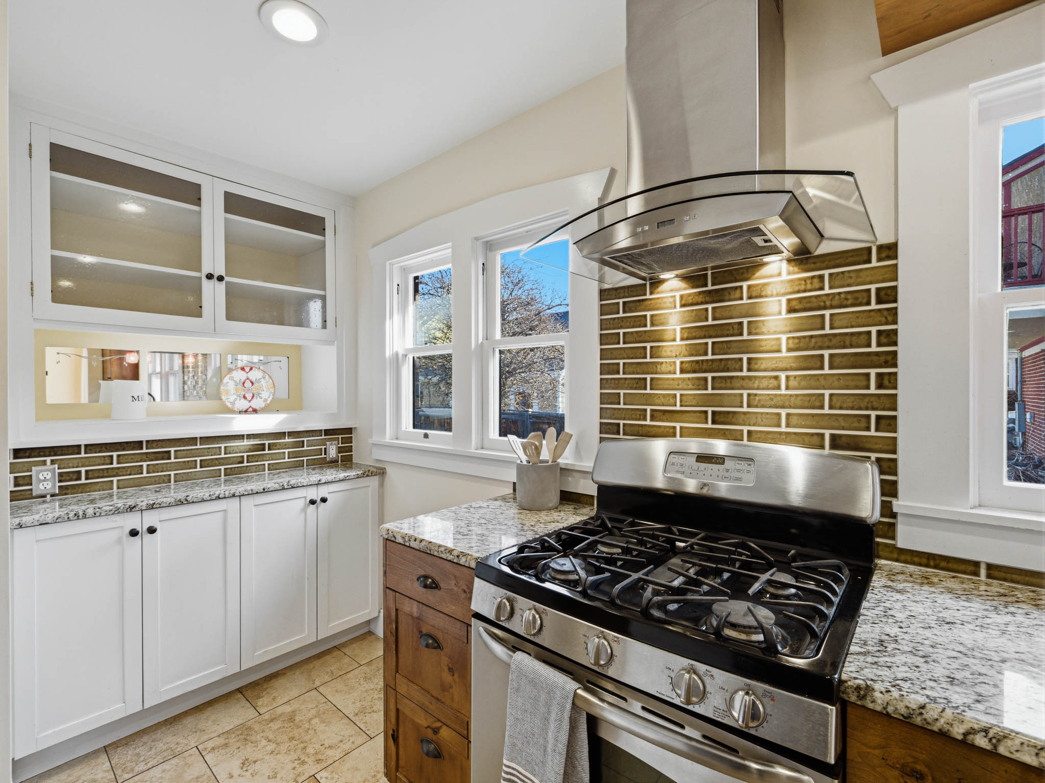 Two tone kitchen with stainless steel gas range oven, two tone cabinetry, glass insert cabinets, light stone counters, and island range hood