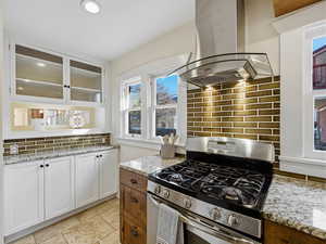 Two tone kitchen with stainless steel gas range oven, two tone cabinetry, glass insert cabinets, light stone counters, and island range hood