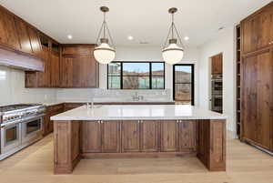 Kitchen with a large island with sink, stainless steel appliances, backsplash, light stone countertops, and light wood-style flooring