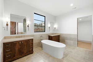 Full bath featuring two vanities, tile walls, a freestanding tub, a wainscoted wall, and recessed lighting
