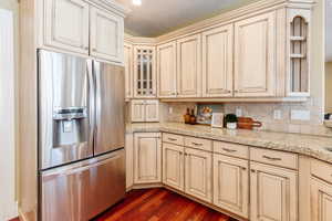 Kitchen with cream cabinets, stainless steel refrigerator with ice dispenser, light stone countertops, and dark wood-type flooring