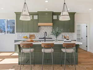 Kitchen featuring green cabinetry, light wood-type flooring, a kitchen bar, a center island with sink, and decorative backsplash