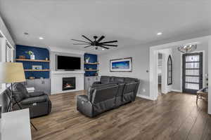 Living room with wood finished floors, a warm lit fireplace, built in shelves, and ceiling fan