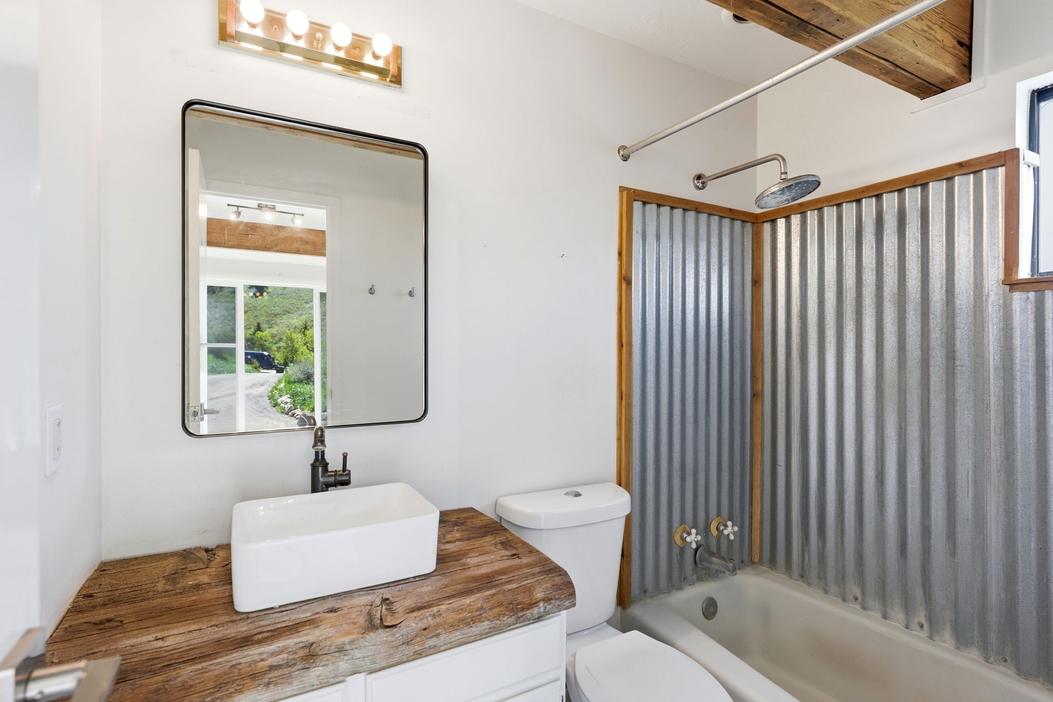 Bathroom featuring a rustic wood vanity with a white vessel sink and matte black faucet, a framed mirror, a four-bulb vanity light fixture, and a corrugated metal shower surround with wood trim