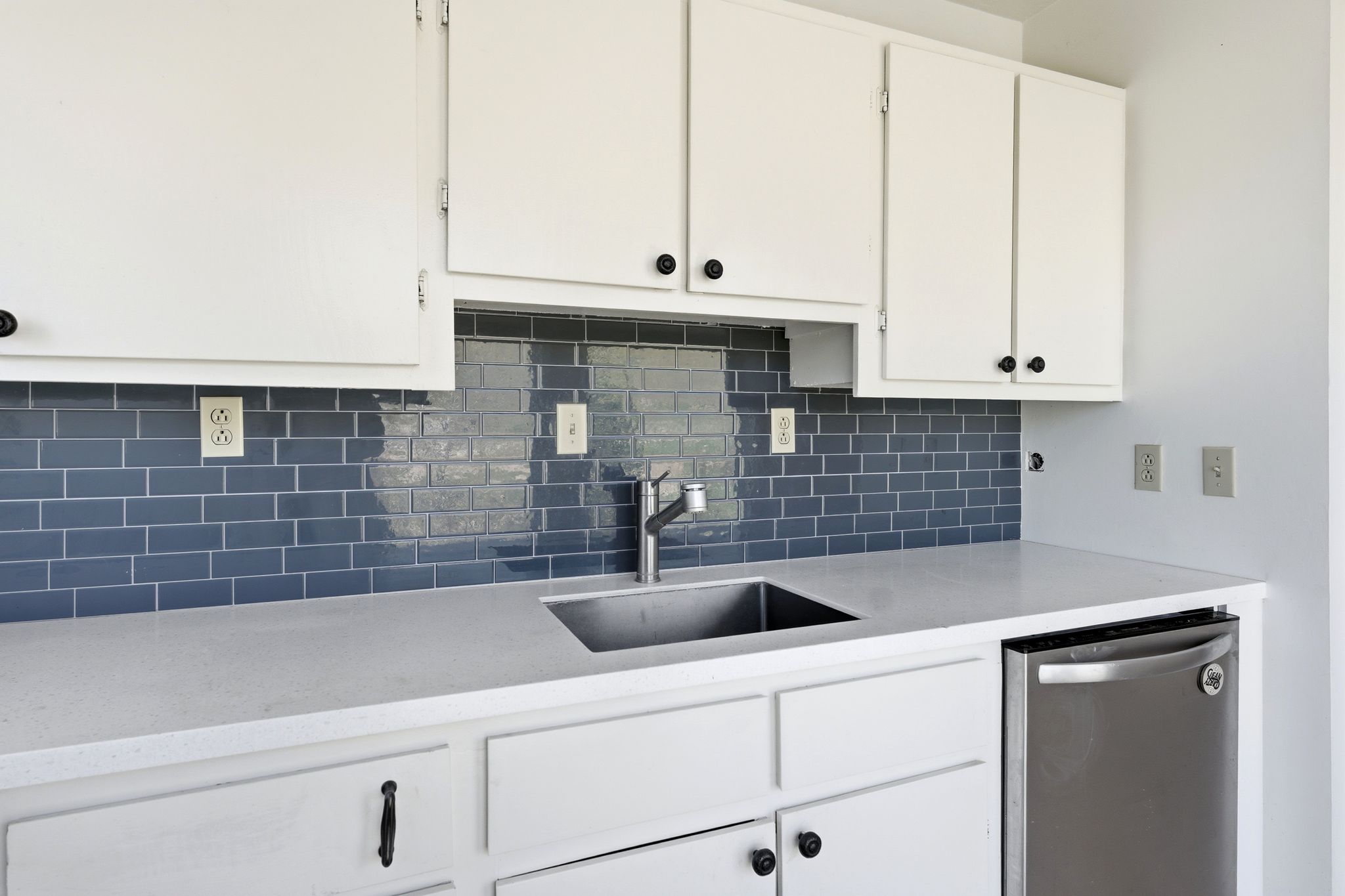 Kitchen featuring white cabinetry, a blue subway tile backsplash, light-colored countertops, a stainless steel sink, and a stainless steel dishwasher
