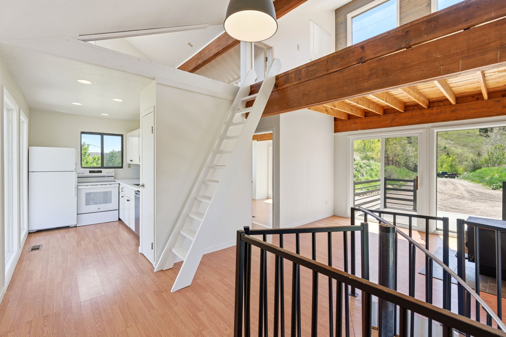 Open-concept living space featuring wood-finish flooring, a functional kitchen with white cabinetry and appliances, a lofted area with exposed wood beams, and a white ladder access