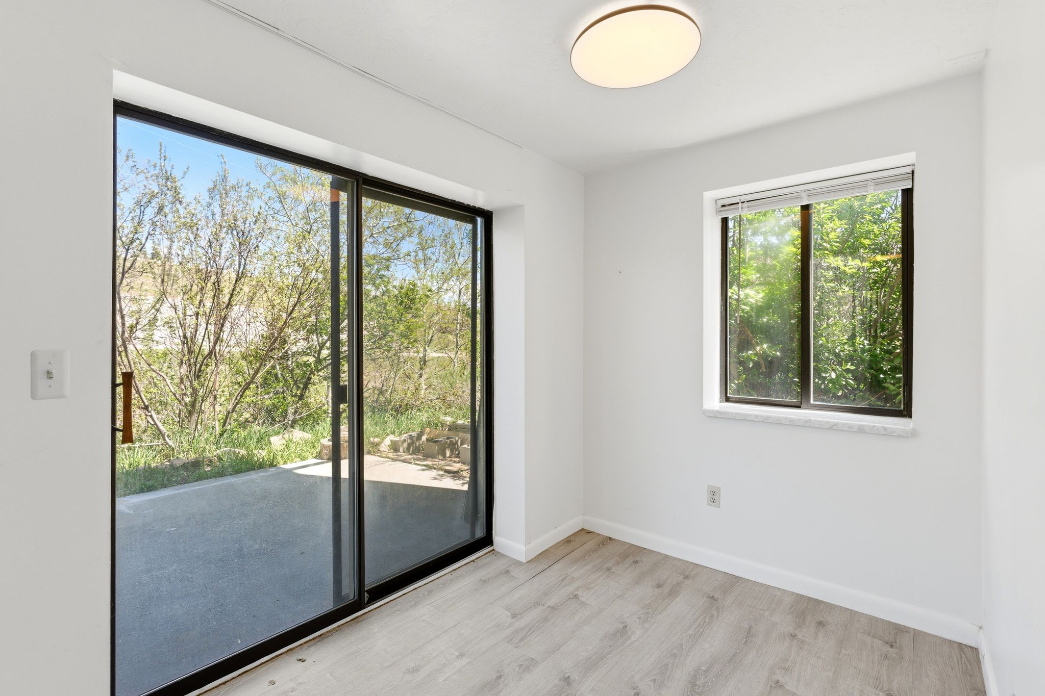 Room featuring a sliding glass door opening to a concrete patio, a rectangular window with green foliage beyond, wood-finish flooring, and a flush-mount ceiling light