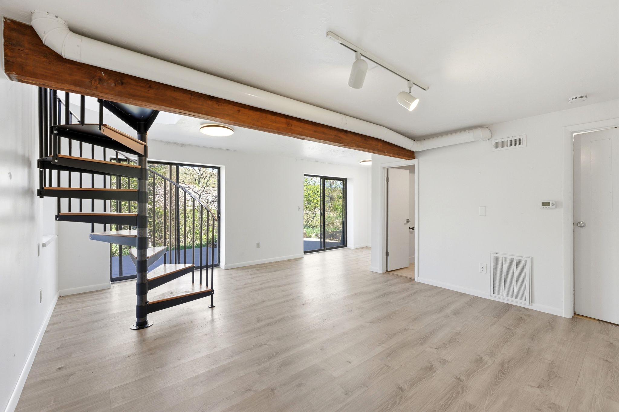 Open concept living area featuring a spiral staircase with wood-finish treads and metal railings, light wood-finish flooring, exposed wood beam, track lighting, and sliding glass doors
