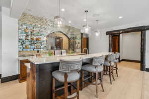 Indoor wet bar with hanging light fixtures, light wood-style flooring, light stone counters, wood finish cabinets, and backsplash